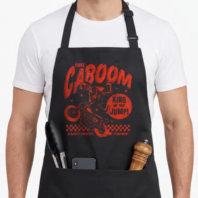 Duke dog breed aprons - Red retro Duke Caboom jumping stuntman with checkered border and bold lettering — shown on a black cotton canvas apron against a white background