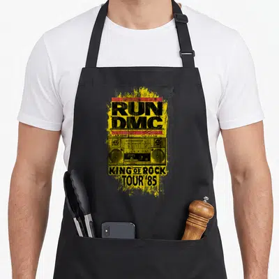 Run king apron - Distressed yellow splatter with boombox and RUN DMC KING OF ROCK TOUR '85 text — shown on a black cotton canvas apron against a white background