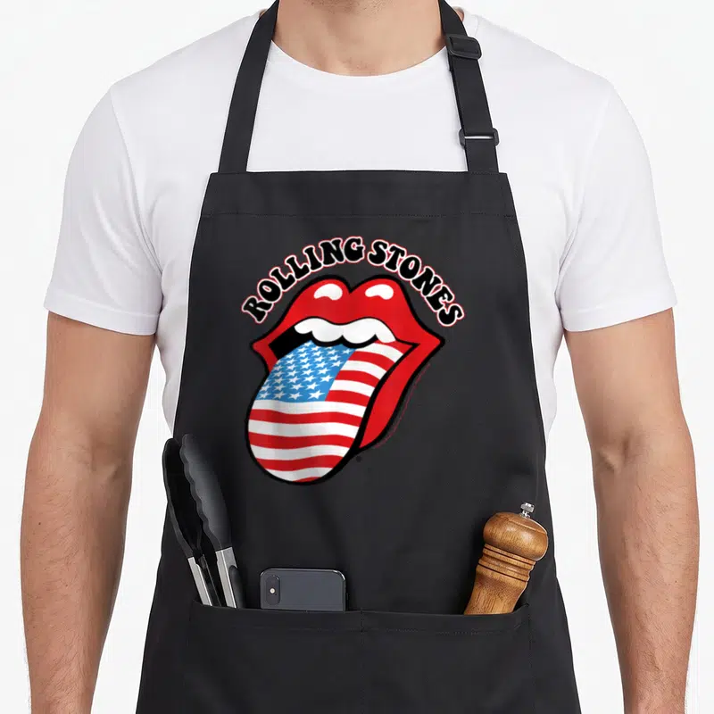 Cute tongue rolling stones apron - ROLLING STONES text over red mouth with USA flag pattern tongue in cartoon style — shown on a black cotton canvas apron against a white background