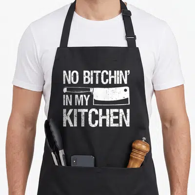 Cute culinary apron - Distressed white text NO BITCHIN IN MY KITCHEN surrounding a meat cleaver on black background — shown on a black cotton canvas apron against a white background