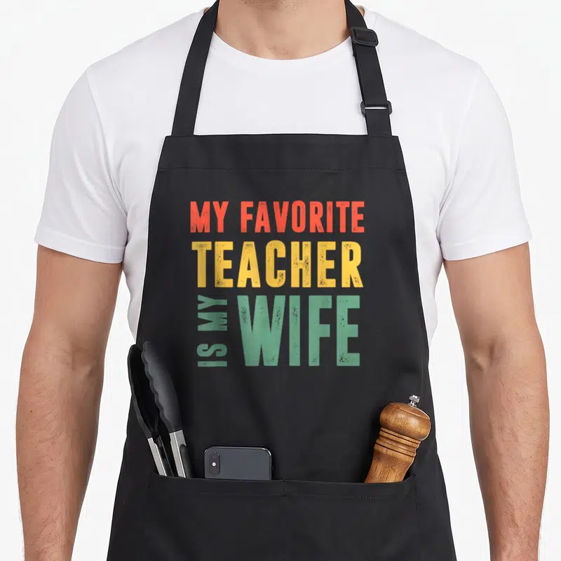 Wisdom teacher pocket apron - Stacked orange red yellow and green text reading My Favorite Teacher Is My Wife — shown on a black cotton canvas apron against a white background