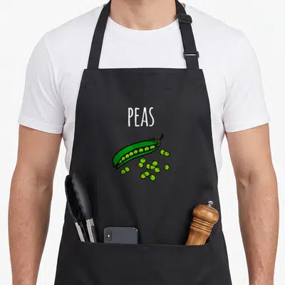 Peas carrot halloween apron - White PEAS text above open green pea pod with scattered round peas in cartoon style — shown on a black cotton canvas apron against a white background