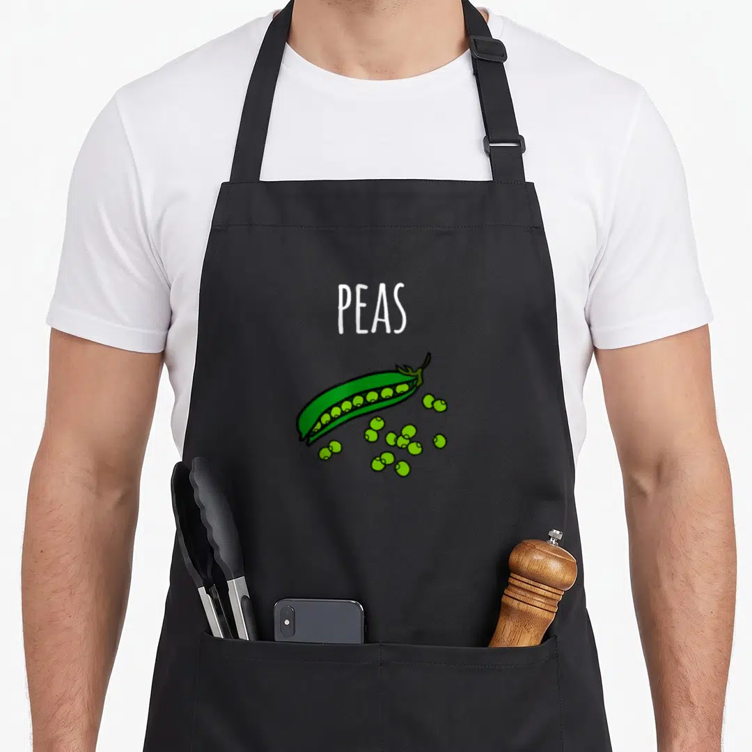 Peas carrot halloween apron - White PEAS text above open green pea pod with scattered round peas in cartoon style — shown on a black cotton canvas apron against a white background