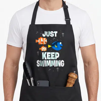 Finding fish apron - Three cartoon fish and jellyfish around Just Keep Swimming text in white blue orange yellow — shown on a black cotton canvas apron against a white background
