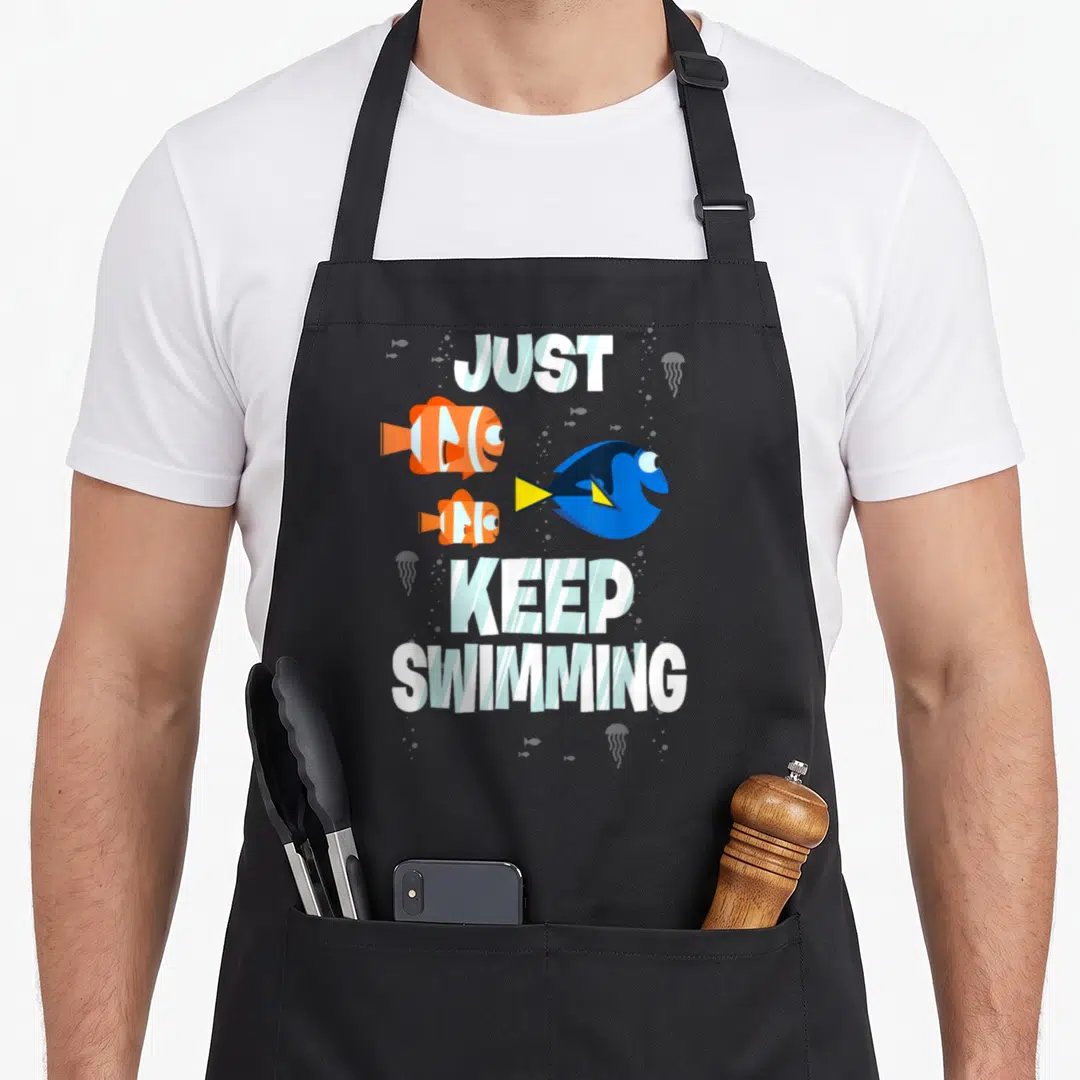 Finding fish apron - Three cartoon fish and jellyfish around Just Keep Swimming text in white blue orange yellow — shown on a black cotton canvas apron against a white background