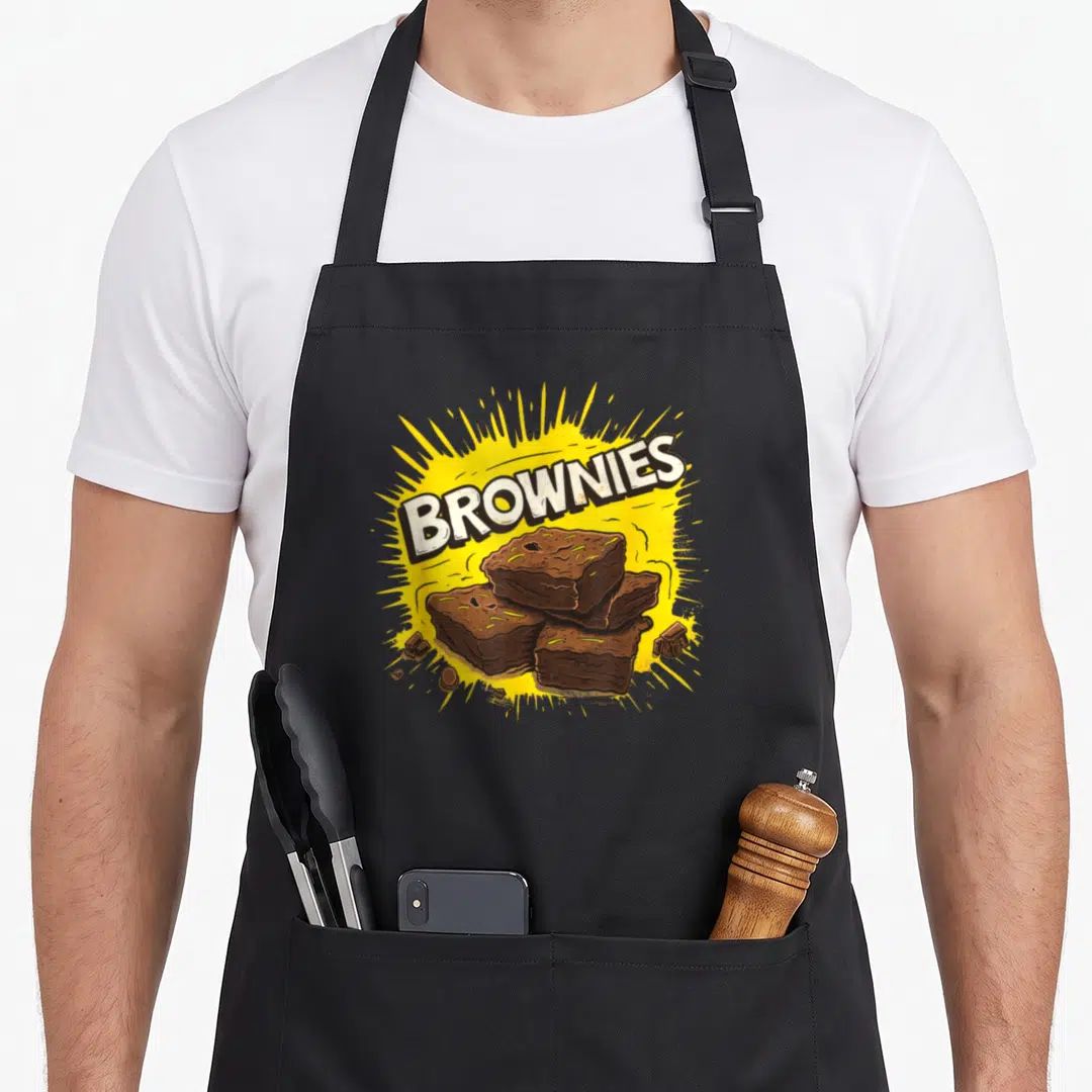 Browny cake apron - BROWNIES text over four stacked brownies on yellow burst with scattered chocolate chunks — shown on a black cotton canvas apron against a white background