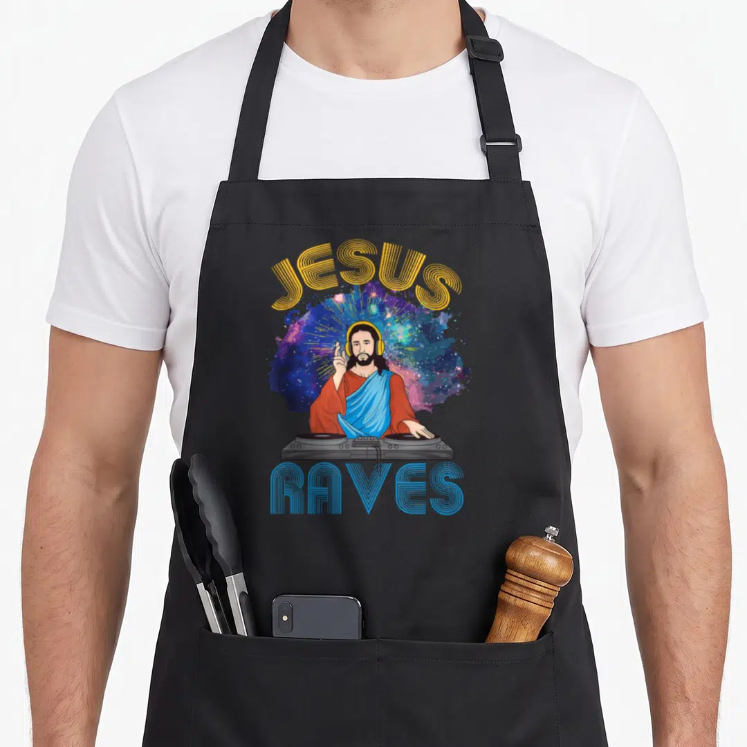 Jesus edm apron - Cartoon Jesus wearing headphones at a DJ deck with arched JESUS RAVES text over a galaxy background — shown on a black cotton canvas apron against a white background