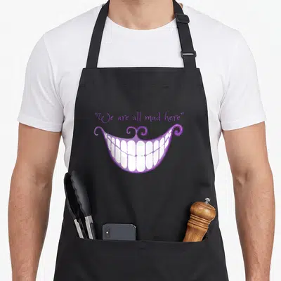 Smile cat apron - Purple cartoon mouth with white teeth and We are all mad here text — shown on a black cotton canvas apron against a white background