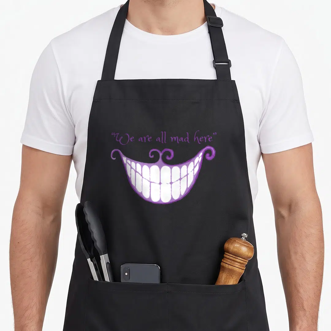 Smile cat apron - Purple cartoon mouth with white teeth and We are all mad here text — shown on a black cotton canvas apron against a white background