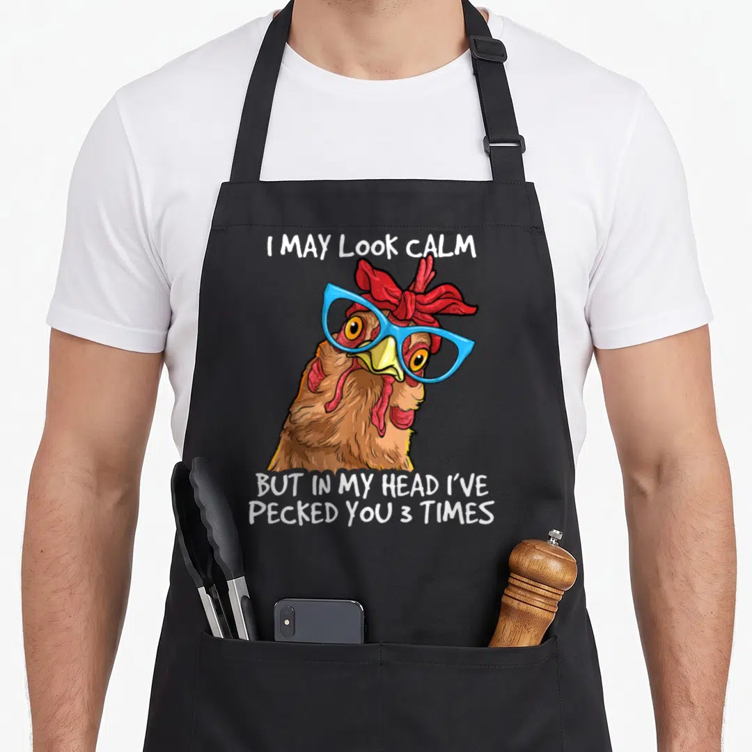 May pecked you apron - Cartoon brown chicken with blue glasses and red headband surrounded by white sarcastic text — shown on a black cotton canvas apron against a white background