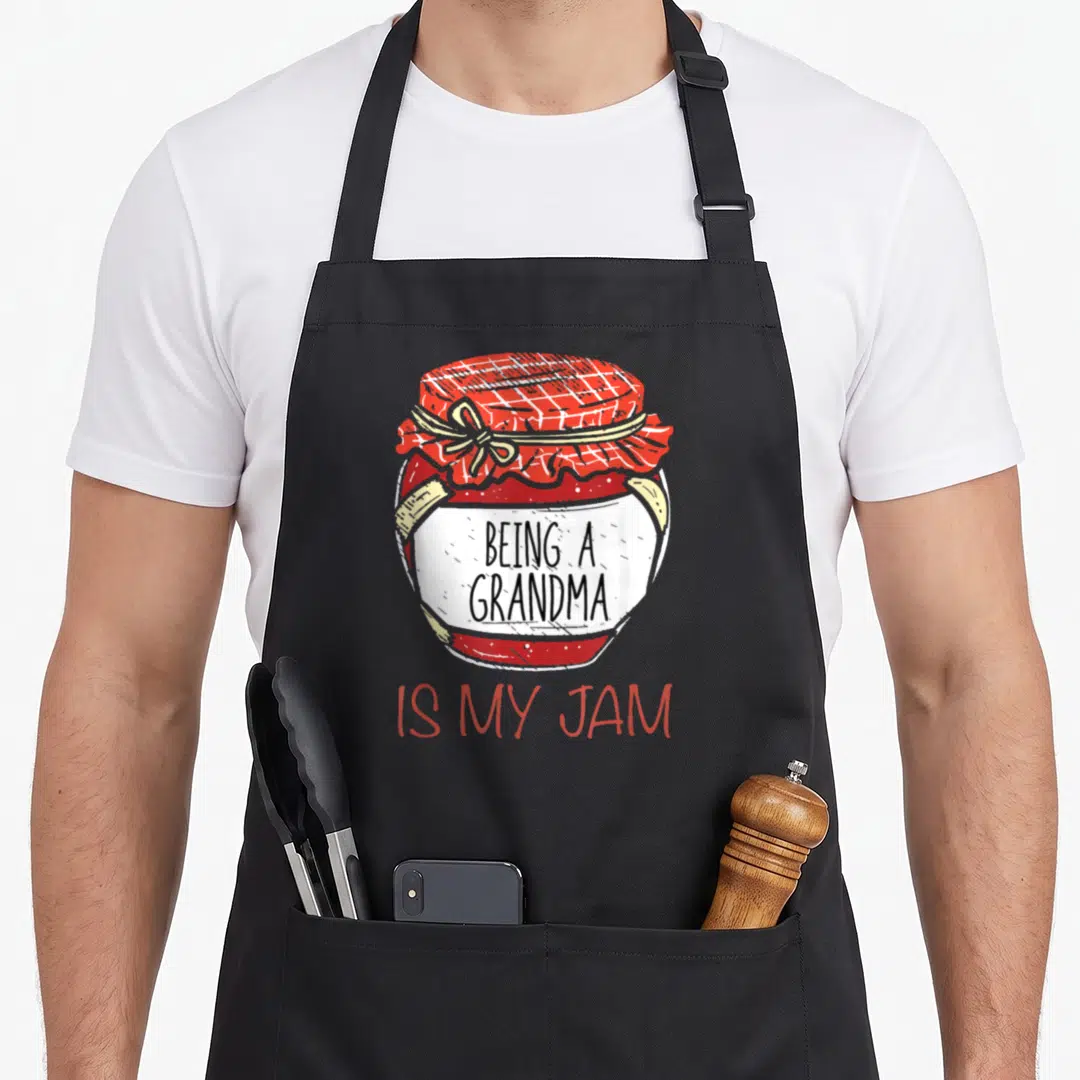 Homemade canning apron - Cartoon red jam jar with white label reading Being a Grandma is my jam in black and red text — shown on a black cotton canvas apron against a white background