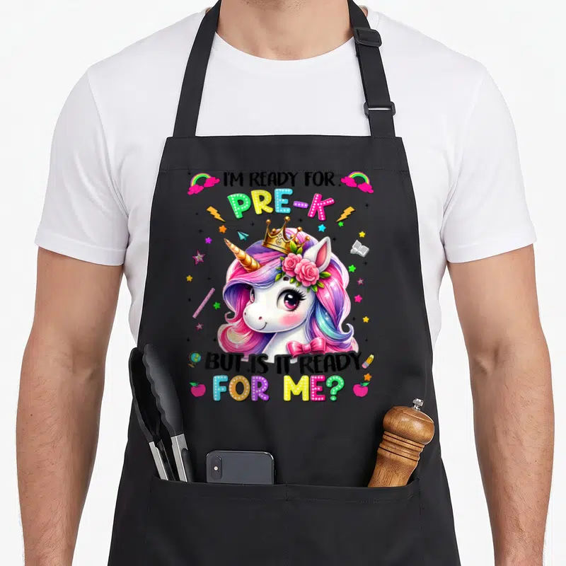 Prek unicorn apron - Cartoon unicorn with crown and roses surrounded by ruler, apple, rainbow, and text — shown on a black cotton canvas apron against a white background