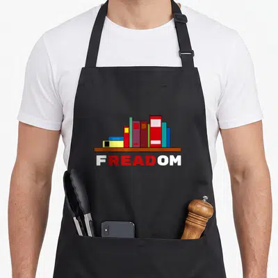 Freadom i love learning apron - Cartoon books on brown shelf with FREADOM text in red and white — shown on a black cotton canvas apron against a white background