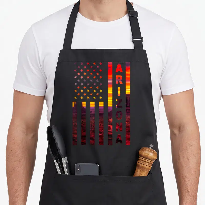 Sunset american flag apron - US flag filled with red orange yellow and purple sunset landscape and vertical ARIZONA text — shown on a black cotton canvas apron against a white background