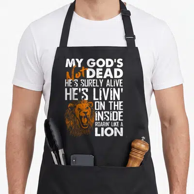 Dead cross apron - My God's Not Dead text stacked with orange lion head overlapping bottom left corner — shown on a black cotton canvas apron against a white background