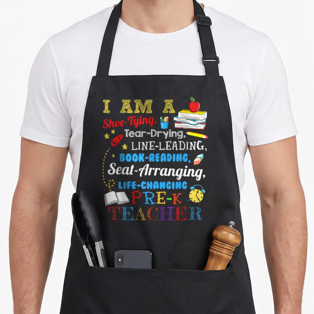 Shoe teacher apron - Stacked text reading I Am A Shoe Tying Teacher surrounded by apple, rocket, books, and alarm clock — shown on a black cotton canvas apron against a white background