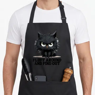 Find cat apron - Grumpy black cat illustration with green eyes above bold text FLUFF AROUND AND FIND OUT — shown on a black cotton canvas apron against a white background