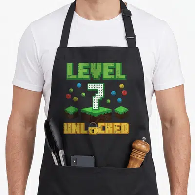Gaming birthday apron - Pixel art LEVEL 7 UNLOCKED text with grass blocks and colorful spheres — shown on a black cotton canvas apron against a white background