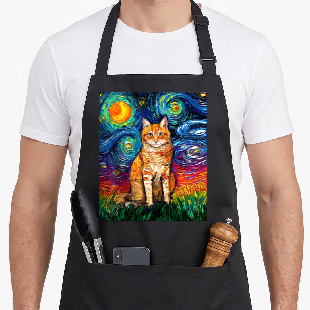 Tabby kitty cat apron - Orange tabby cat sitting on green grass against a swirling blue and yellow starry night — shown on a black cotton canvas apron against a white background