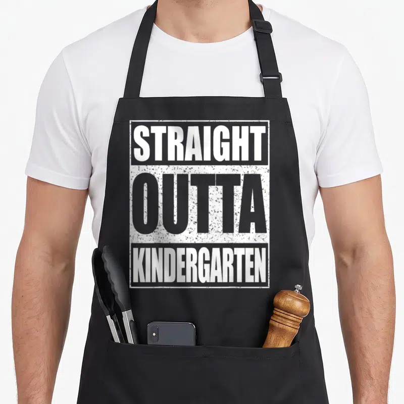 Outta straight a student apron - Stacked white and grey text reading STRAIGHT OUTTA KINDERGARTEN inside a distressed — shown on a black cotton canvas apron against a white background
