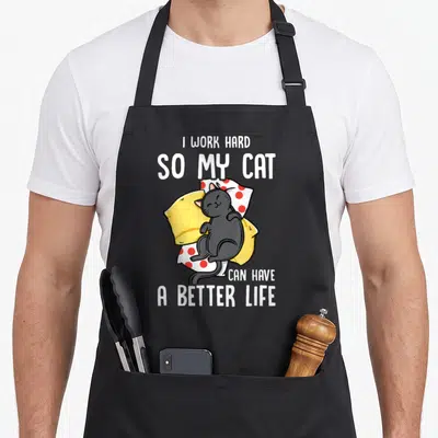 Hard cat apron - Cartoon black cat sleeping on yellow pillow and red polka dot blanket with white text — shown on a black cotton canvas apron against a white background