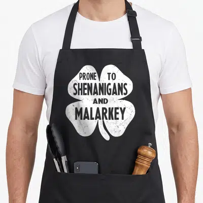 Prone st patrick day aprons - White distressed four-leaf clover with stacked text PRONE TO SHENANIGANS AND MALARKEY — shown on a black cotton canvas apron against a white background
