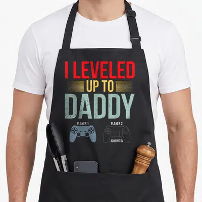 1st daddy apron - I leveled up to daddy text with solid blue and white outlined game controllers — shown on a black cotton canvas apron against a white background