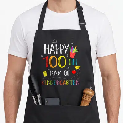 School kindergarten teacher apron - Happy 100th Day of Kindergarten text with pencils, apple, plane, and notebook — shown on a black cotton canvas apron against a white background