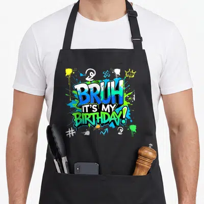 Bruh colorful aprons - Graffiti text BRUH IT'S MY BIRTHDAY with blue green yellow paint splatters and crown symbol — shown on a black cotton canvas apron against a white background
