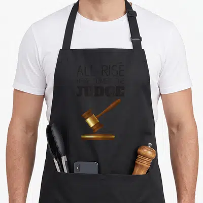 Rise come and get it apron - All Rise Here Comes The Judge text above a brown and gold gavel on a sound block — shown on a black cotton canvas apron against a white background