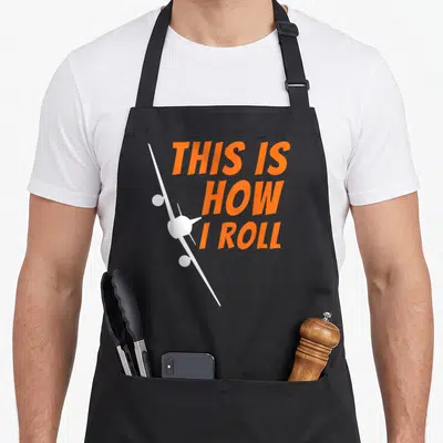 Roll pilot aviation airplane apron - White airplane silhouette overlapping orange stacked text reading THIS IS HOW I — shown on a black cotton canvas apron against a white background