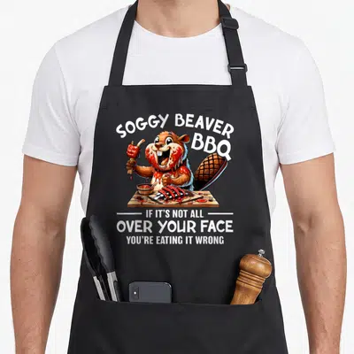 Face beaver apron - Cartoon beaver holding rib with sauce and text reading Soggy Beaver BBQ If Its Not All Over Your — shown on a black cotton canvas apron against a white background