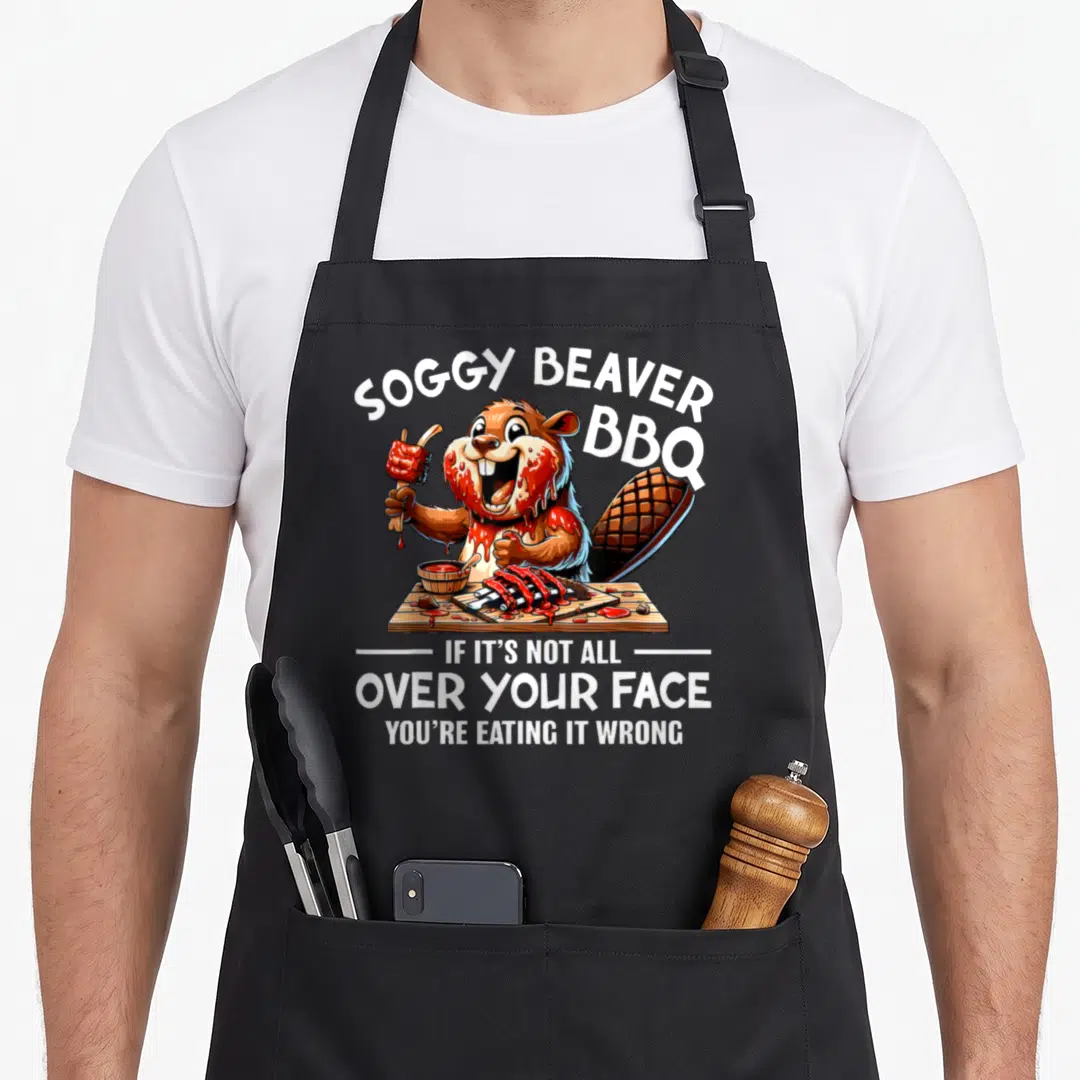 Face beaver apron - Cartoon beaver holding rib with sauce and text reading Soggy Beaver BBQ If Its Not All Over Your — shown on a black cotton canvas apron against a white background