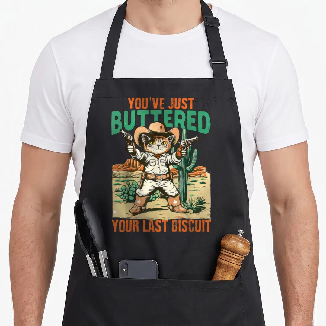 Cowboy cat apron - Cartoon cat in cowboy hat holding guns between cacti with orange and green text — shown on a black cotton canvas apron against a white background