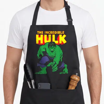 Incredible distressed apron - Crouching green figure with purple pants below stacked THE INCREDIBLE HULK text in yellow — shown on a black cotton canvas apron against a white background