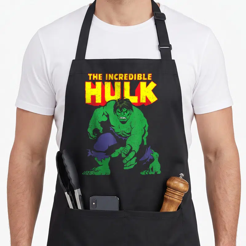 Incredible distressed apron - Crouching green figure with purple pants below stacked THE INCREDIBLE HULK text in yellow — shown on a black cotton canvas apron against a white background