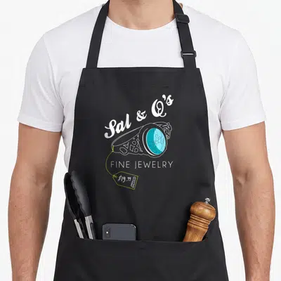 Impractical jewelry apron - White line-art ring with blue gem and yellow price tag reading Sal and Qs Fine Jewelry — shown on a black cotton canvas apron against a white background