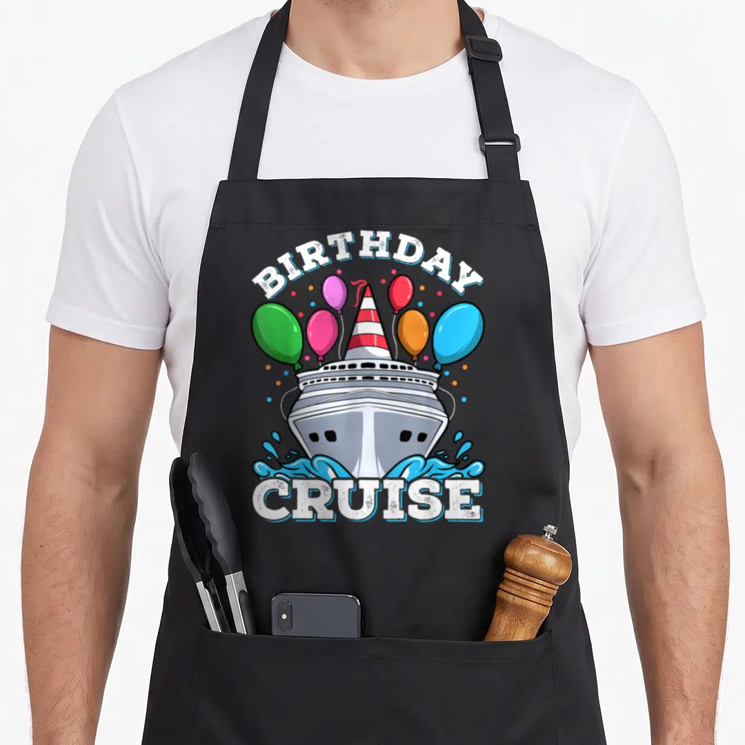 Cruising birthday apron - Cartoon ship wearing party hat surrounded by balloons and BIRTHDAY CRUISE text — shown on a black cotton canvas apron against a white background