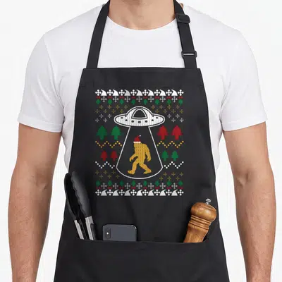 Ufo santa claus apron - Golden Bigfoot in red Santa hat inside white UFO beam surrounded by green red trees and white — shown on a black cotton canvas apron against a white background