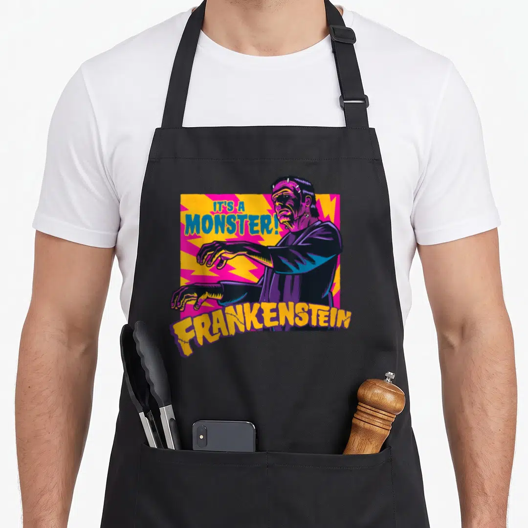 Monster frankenstein apron - Cartoon Frankenstein monster in purple and blue with yellow text IT'S A MONSTER — shown on a black cotton canvas apron against a white background