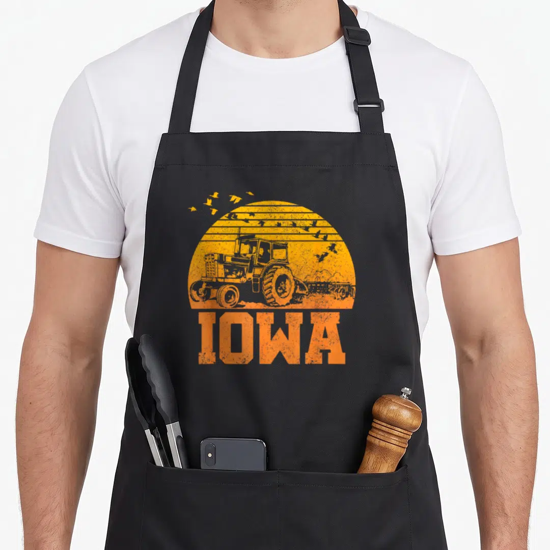 Iowa farmer apron - Retro tractor silhouette inside orange sun with flying birds and IOWA text below — shown on a black cotton canvas apron against a white background