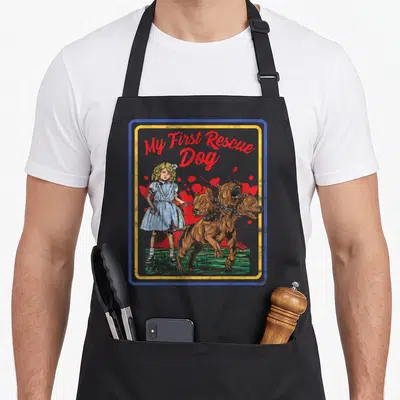 Game grill rescue apron - Girl holding leash attached to brown three-headed dog under red My First Rescue Dog text — shown on a black cotton canvas apron against a white background