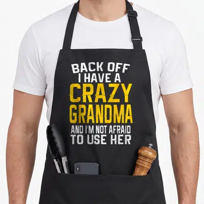 Retro grandmother grandma apron - White and yellow distressed text reading BACK OFF I HAVE A CRAZY GRANDMA AND I'M NOT — shown on a black cotton canvas apron against a white background