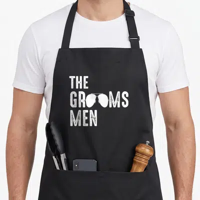 Shade groom apron - White distressed text reading The Grooms Men with sunglasses replacing O's in middle line — shown on a black cotton canvas apron against a white background