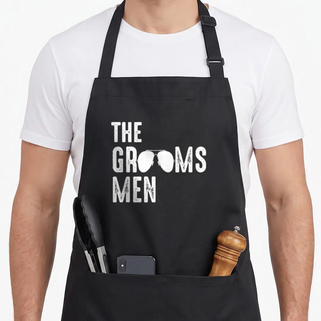 Shade groom apron - White distressed text reading The Grooms Men with sunglasses replacing O's in middle line — shown on a black cotton canvas apron against a white background