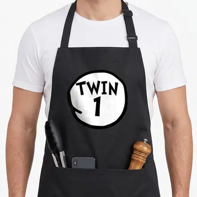 Funny best twin ever apron - White circle with thick black border containing TWIN 1 text — shown on a black cotton canvas apron against a white background