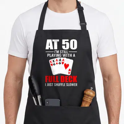 Poker birthday apron - AT 50 text with fan of five playing cards KKAA Ace — shown on a black cotton canvas apron against a white background