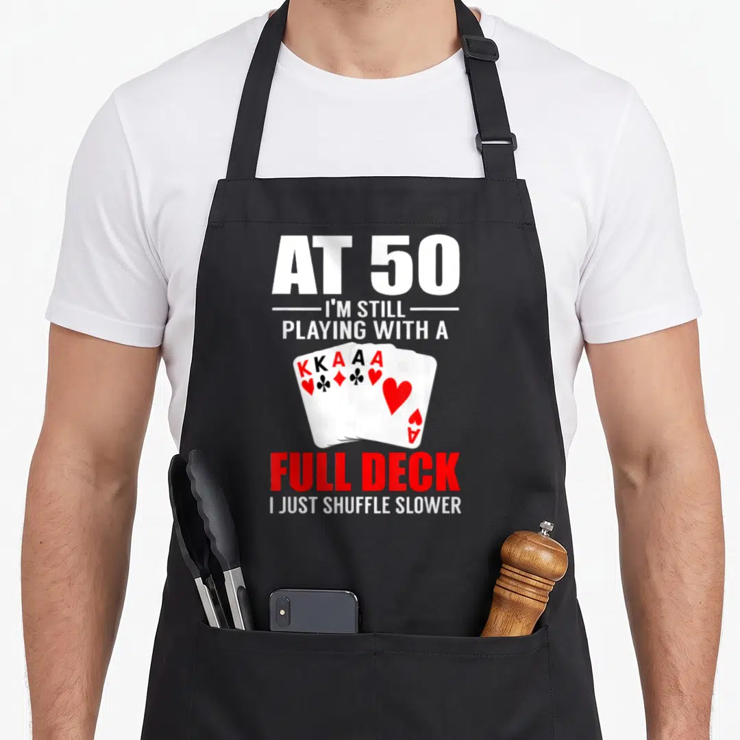 Poker birthday apron - AT 50 text with fan of five playing cards KKAA Ace — shown on a black cotton canvas apron against a white background