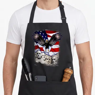 Mount 4th of july apron - Realistic bald eagle over red white blue flag and grey Mount Rushmore faces — shown on a black cotton canvas apron against a white background