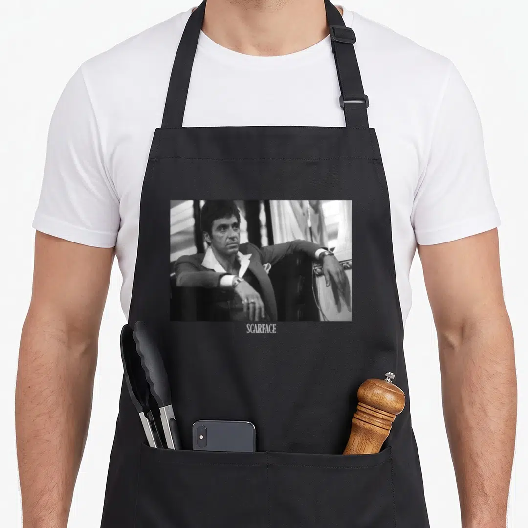 Tony scaredy apron - Grayscale photo of Tony Montana sitting in a chair with SCARFACE text below — shown on a black cotton canvas apron against a white background